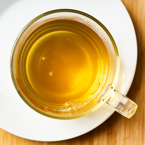 Taza de vidrio transparente llena de té verde humeante en un platillo blanco.