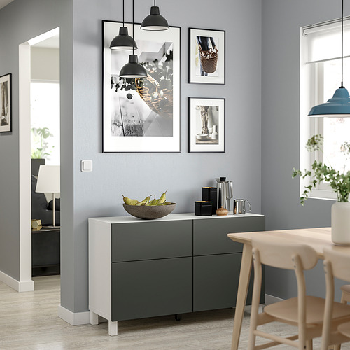 Sala de estar moderna con un mueble de almacenamiento BESTÅ blanco y gris, elegantes lámparas colgantes, fotos enmarcadas y una mesa de madera con sillas.