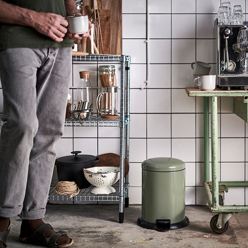 Persona sosteniendo una taza en una cocina acogedora con pared de azulejos, estante de metal y bote de basura.