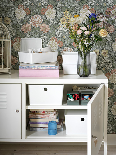 Armario metálico blanco IKEA PS; papel pintado floral; arriba: cajas de almacenamiento, libros, jarrón con flores; Inferior: puerta abierta que revela libros, contenedores.