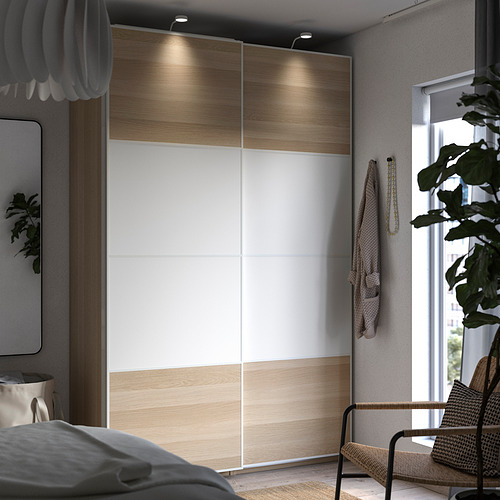 Dormitorio moderno con armario PAX, puertas correderas e iluminación integrada. Silla de ratán y planta junto a ventana.