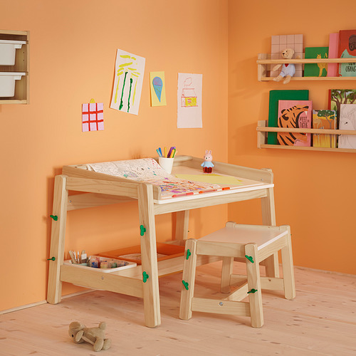 Mesa infantil de madera con altura regulable. Tiene almacenes para materiales de arte, un conejo de juguete y dibujos en la pared.