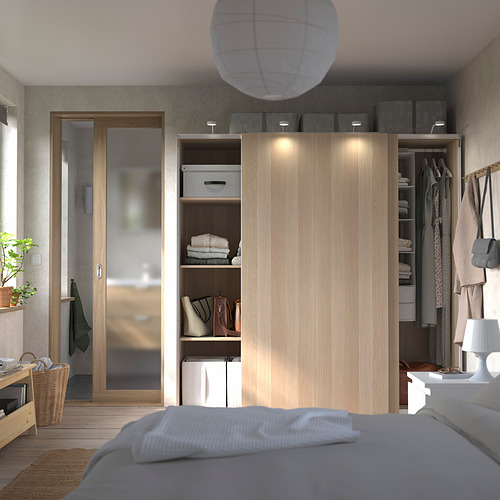 Dormitorio con armario de madera PAX. Tiene puertas correderas, estantes con ropa, bolsos y cajas. Cama gris con manta blanca en primer plano.