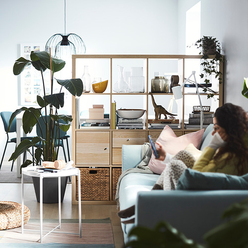Sala de estar moderna con sofá azul claro, persona leyendo, planta, estante KALLAX, luz de ventana.