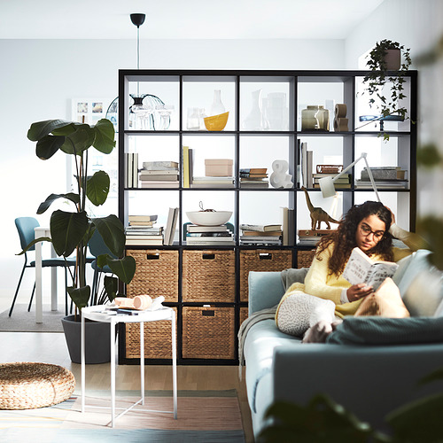 Mujer lee un libro en un sofá azul cerca de un estante KALLAX negro con cestas de mimbre, plantas y decoración.