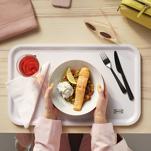 Persona sosteniendo un cuenco blanco de IKEA 365+ con comida. La bandeja contiene cubiertos, una servilleta y una bebida. La ambientación incluye accesorios coloridos.