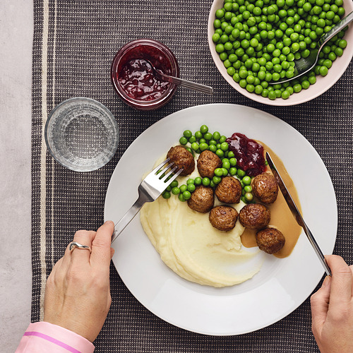 Persona sosteniendo un plato con albóndigas, puré de papas, guisantes y mermelada. Cuencos y tazas rodean el plato.