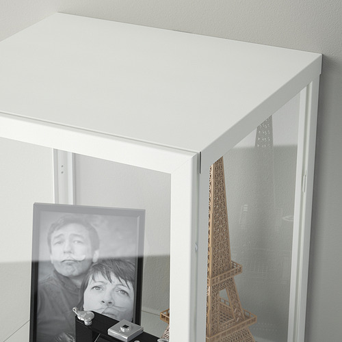 Mueble blanco con puerta de cristal que exhibe artículos. Los estantes sostienen una foto enmarcada y un modelo de torre Eiffel, que muestran las funciones de almacenamiento y exhibición.
