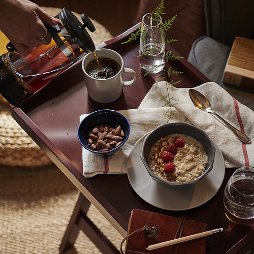 Persona sirviendo café cerca de los artículos del desayuno, incluyendo avena con bayas y almendras en tazones, sobre una mesa con un cuaderno y un lápiz.