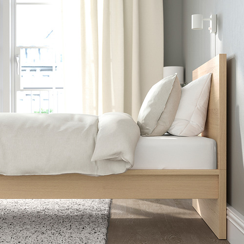 Escena de dormitorio con estructura de cama de madera clara, ropa de cama blanca y almohadas grises. La cabecera se apoya en la pared, cerca de una ventana.