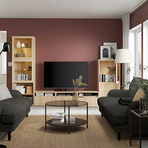 Sala de estar moderna con mueble de televisión BESTÅ, sofás negros, mesa de centro de cristal y luz natural por la ventana.