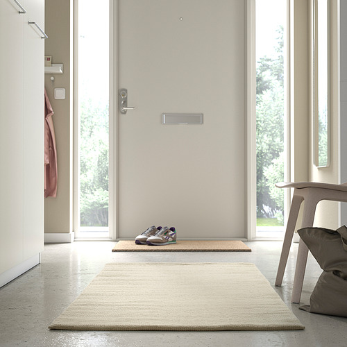 Habitación moderna con alfombra TIDTABELL beige cerca de la puerta, alfombra más pequeña y silla con cojín.