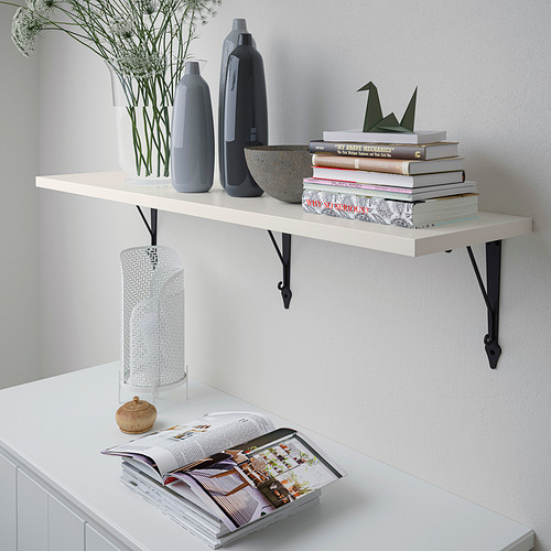 El estante de pared blanco contiene jarrones, libros y una lámpara. Montado con soportes negros.