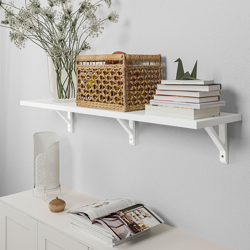 Un estante de pared blanco sostiene una cesta tejida, libros y un jarrón con flores. Está soportado por soportes metálicos.