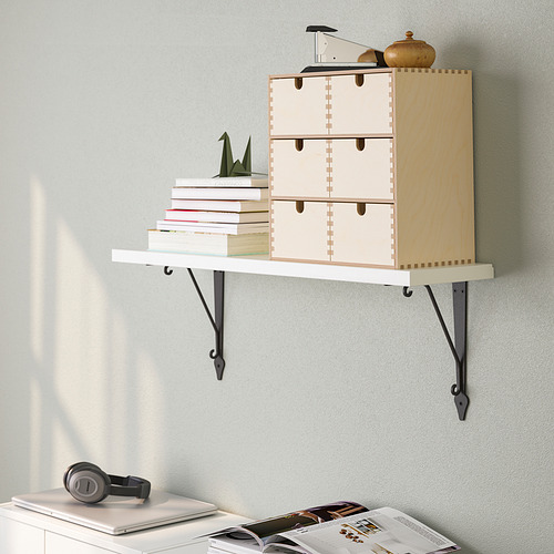 Una estantería de pared con cajones que contienen libros y un elemento decorativo. Abajo, un escritorio con auriculares, una computadora portátil y revistas.