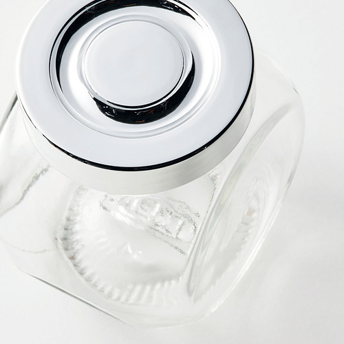 Frasco de vidrio transparente con tapa metálica, que refleja la luz, se asienta sobre una superficie blanca.