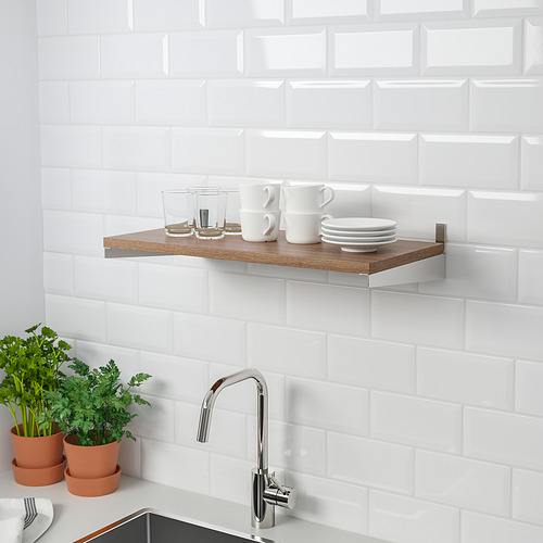 Estantería de cocina con tazas, vasos y platos, montada en una pared de azulejos. Dos hierbas en macetas se encuentran en un mostrador debajo de un grifo cromado.