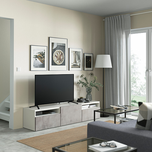 Sala de estar moderna con banco de televisión BESTÅ, TV de pantalla plana, lámpara, arte, mesa de centro de vidrio y planta. Decoración gris con cortinas y alfombra.