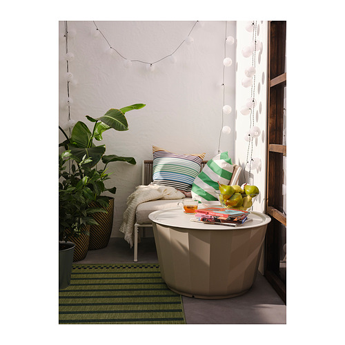 Moderno rincón de exterior con mesa LÅGASKÄR blanca, cojines a rayas, alfombra verde, planta grande y luces colgantes.