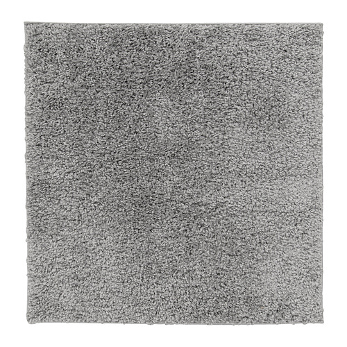 Una alfombra cuadrada gris ÄRENDE con textura peluda, fabricada en polipropileno y caucho sintético, ideal para el confort y la absorción del sonido.