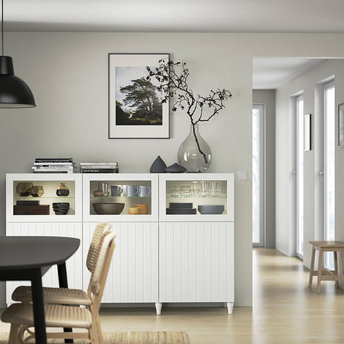 Aparador blanco BESTÅ con puertas de cristal, platos, cuencos, jarrones, arte, mesa de madera, sillas, pasillo.