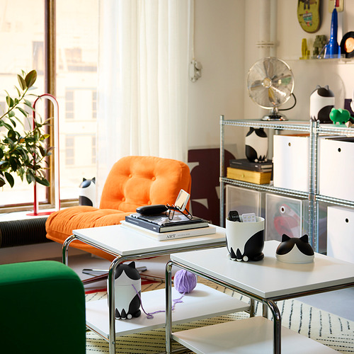 Lindos organizadores con forma de gato en la mesa, uno con libros, otro con control remoto. Silla naranja, cojín verde, hilo cerca.