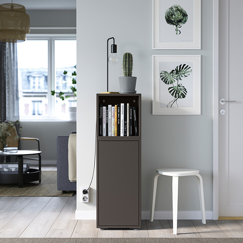 Unidad de almacenamiento EKET gris con puerta y estantes, entre arte vegetal, taburete y cactus.