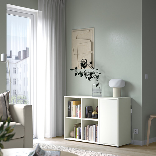 Pequeños estantes de cubos blancos EKET debajo de una ventana, con libros y decoración. Pared azul claro con arte abstracto.
