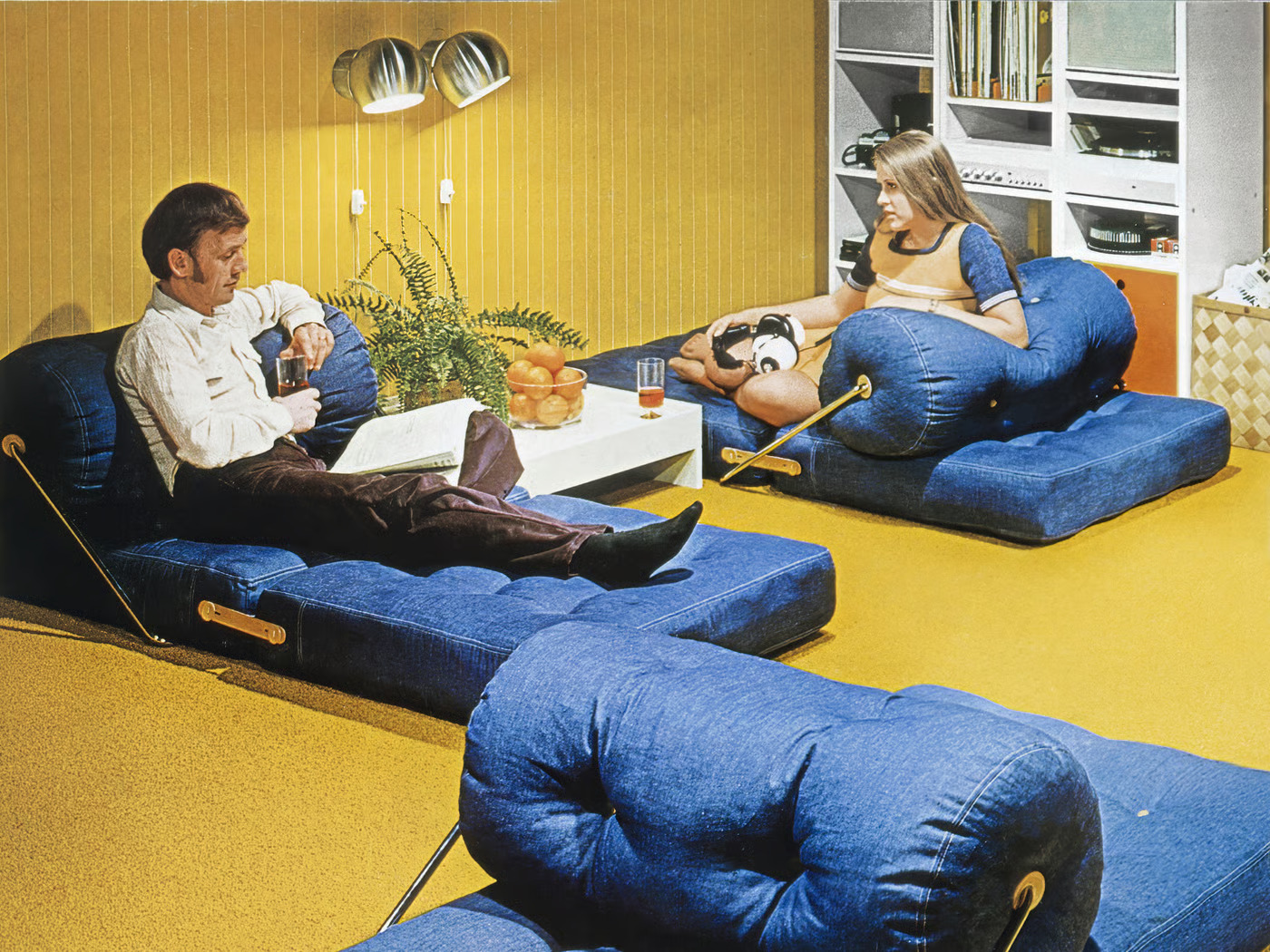 A couple relaxing in a 1970s living room featuring blue denim furniture, yellow walls, and mustard carpeting.
