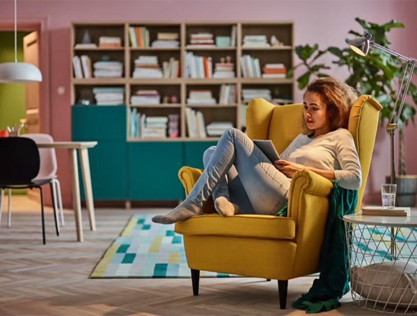 A woman relaxing in a bright yellow wingback chair while using a tablet in a modern, colorful living room.