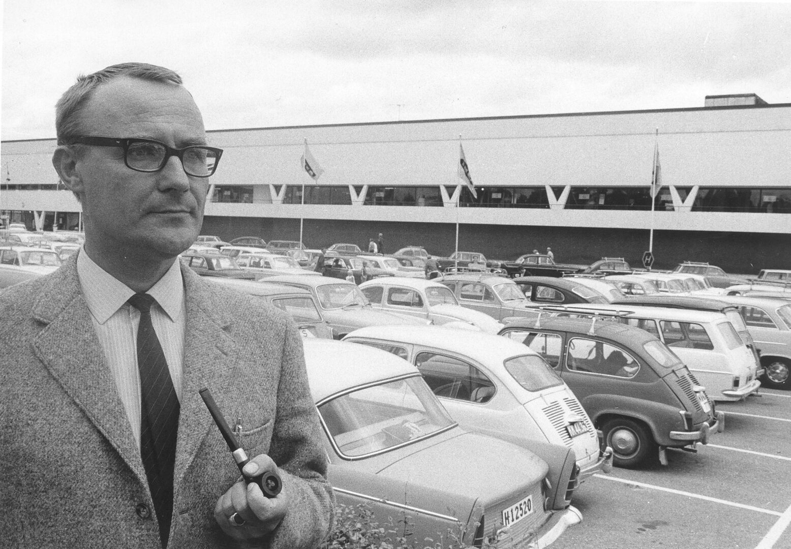 A black and white vintage photo of a man in a suit holding a pipe, with a crowded parking lot of mid-century cars and a long building in the background.
