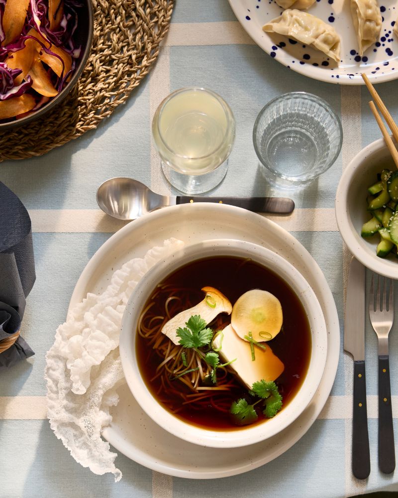 A dotted/light beige DAGGASTER plate and bowl has a soup made of mushrooms, tofu and noodles, next to a bowl of avocados.