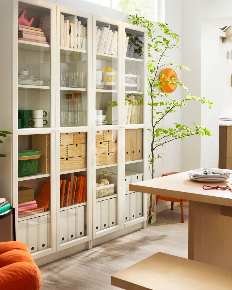 White BILLY bookcases with glass doors, placed next to a table, have binders, boxes, glasses and mugs placed on the shelves.
