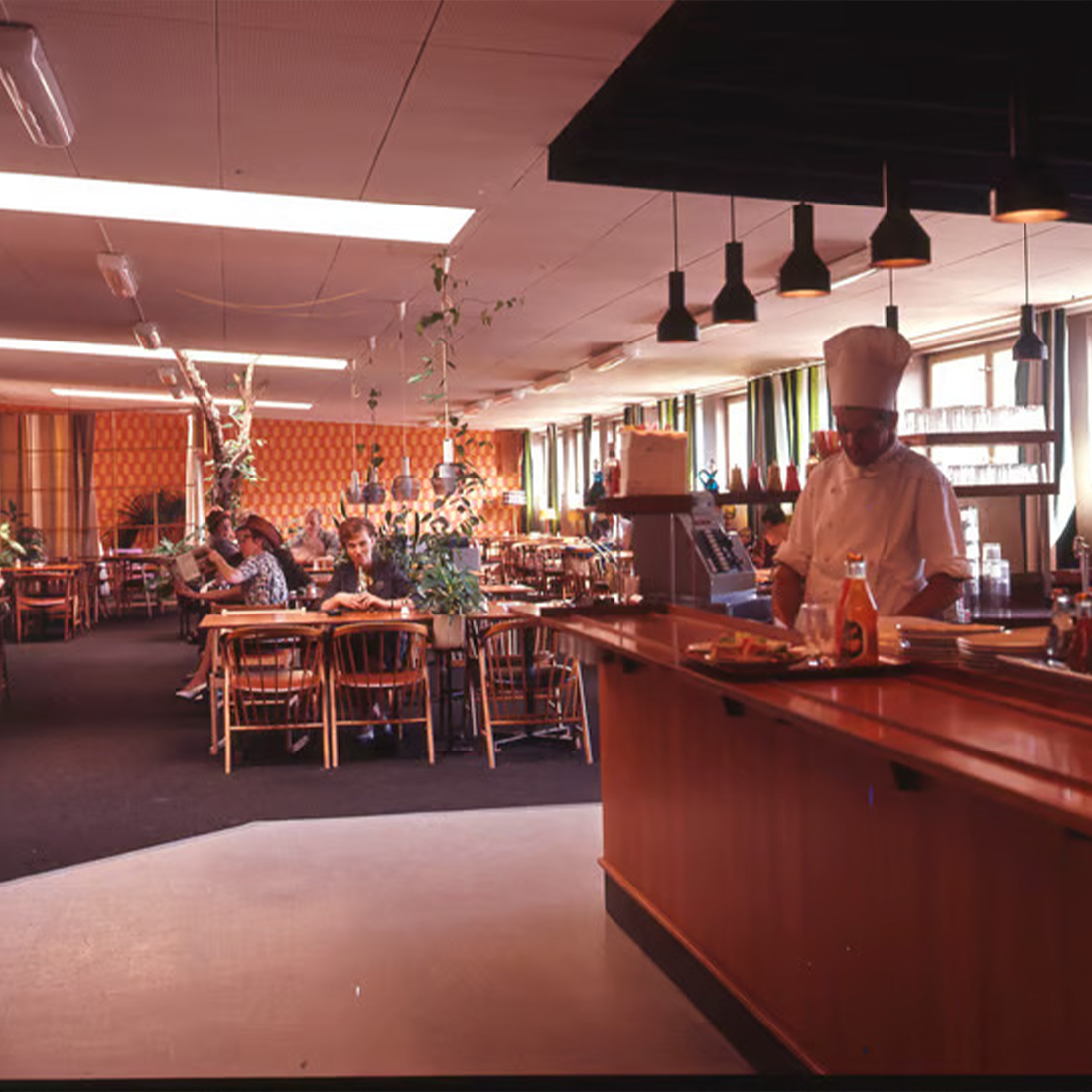 A vintage photograph of a mid-century restaurant interior featuring patrons seated at tables, a chef behind a wooden counter, and retro decor with patterned walls.