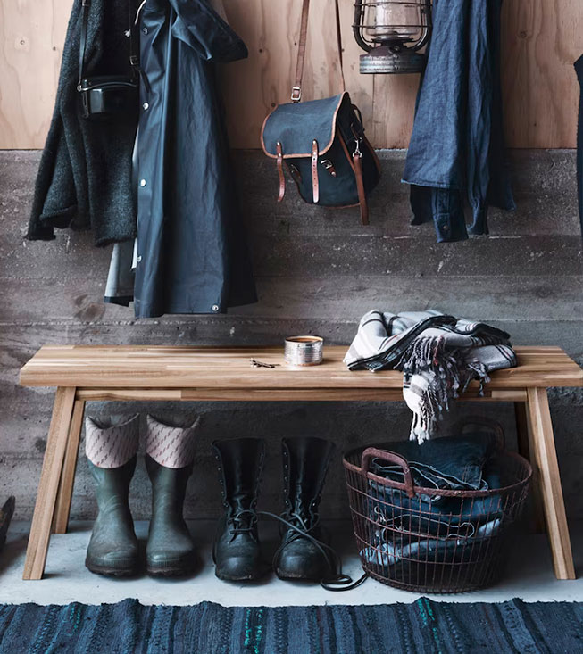 A wooden bench in a rustic entryway, with coats hanging, boots below, and a metal basket.