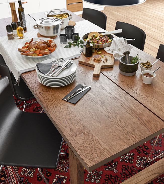 A wooden dining table set with food, plates, and cutlery, over a red rug.
