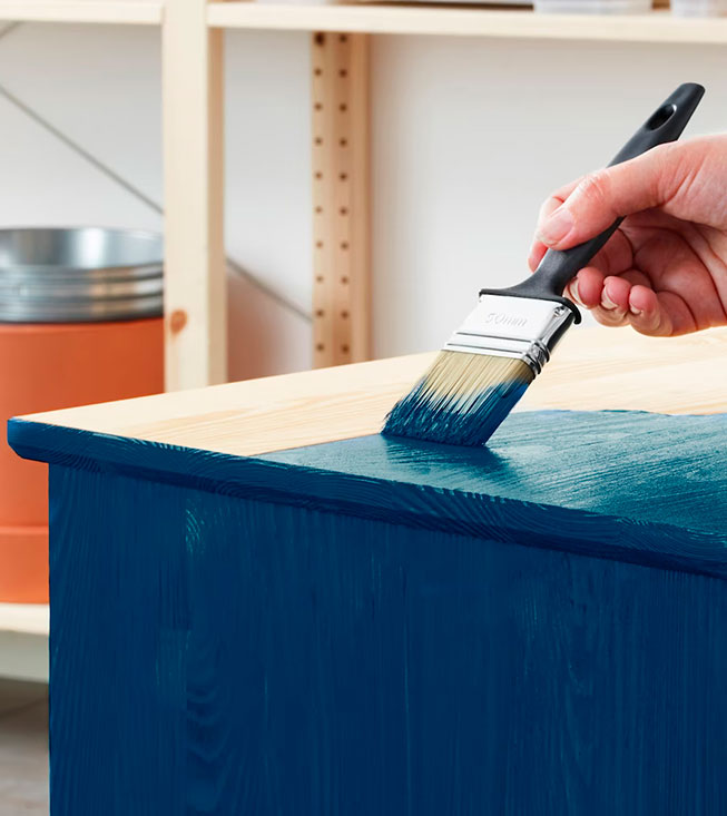A hand paints a wooden surface dark blue with a brush, showing the before and after.