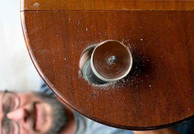A drill bit bores into a wooden surface, with sawdust. Blurred man's face in the bottom left.