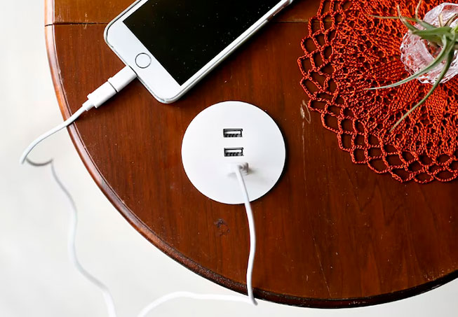 A white phone charging from a round USB hub on a wooden table. Orange doily and plant.