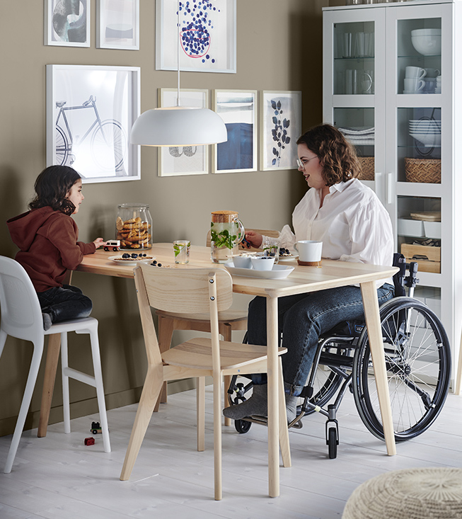 A woman in a wheelchair and a child sit at a light wooden dining table in a bright room.
