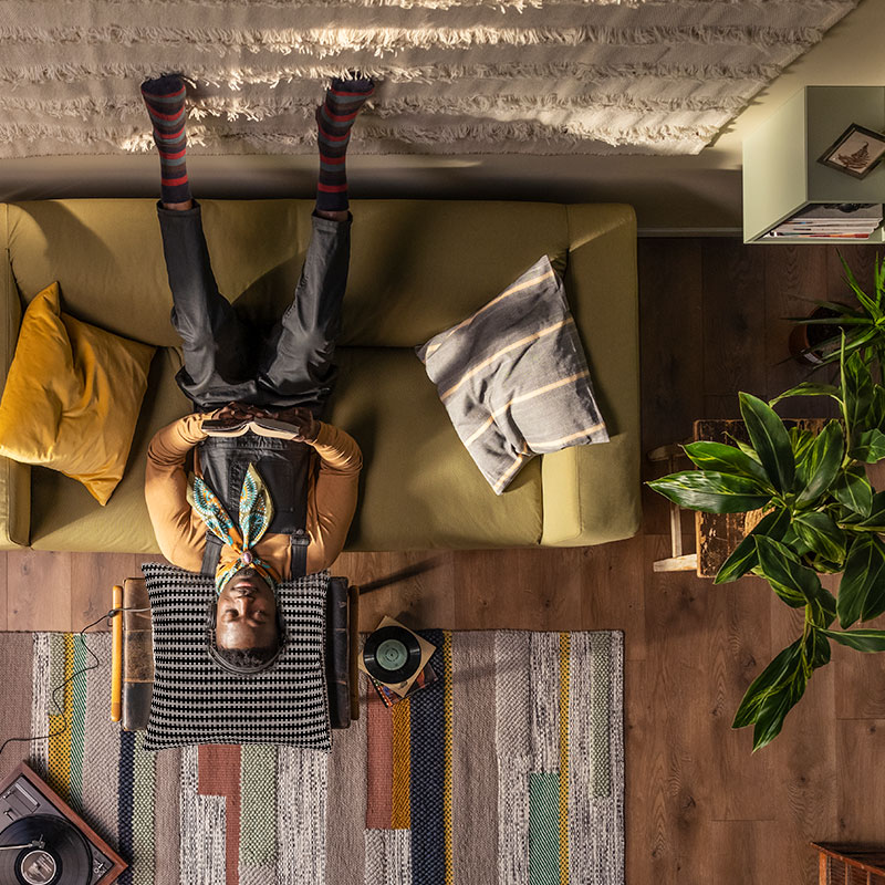 Overhead view: person with bunny ears, upside down on green sofa, cozy living room with plants, guitar, and record player.