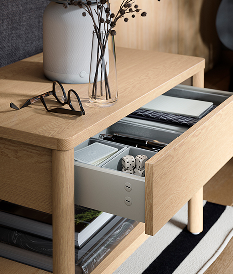 Light wood side table with open drawer showing organizers with small items, next to a decorative vase and a pair of glasses.