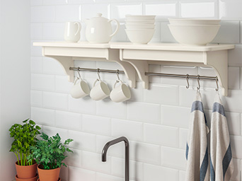 Dos estantes de pared en una cocina, con paños de cocina, tazas, cuencos y una tetera en colores claros; debajo hay un fregadero con grifo.