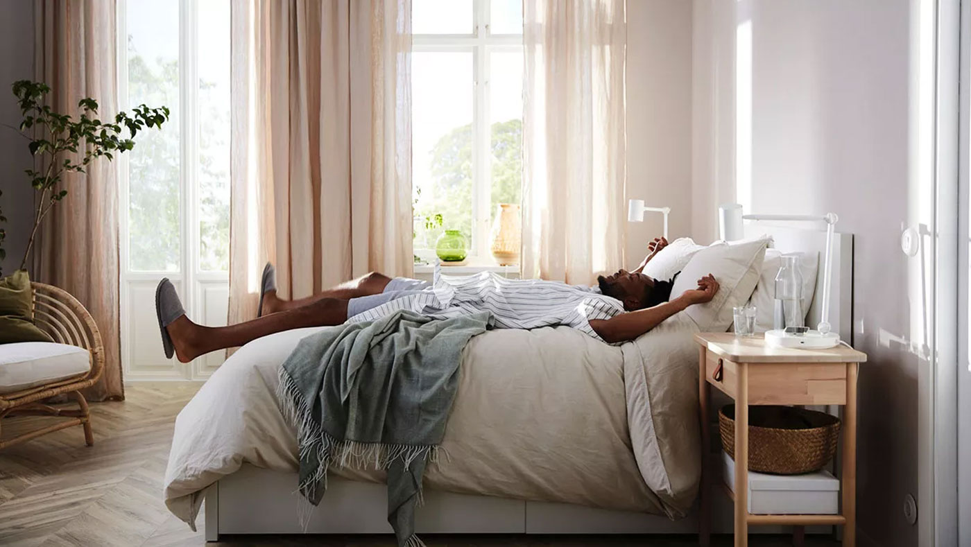 Image of a brightly lit bedroom with beige curtains in side view, with a man lying down as if he just jumped in and sinks between sheets and fluffy bedspreads, the bed has a pine look and modern style.