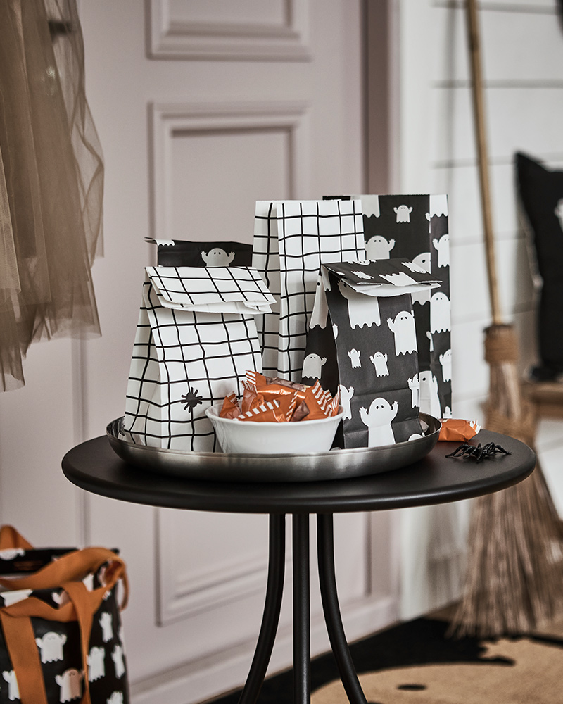 Some KUSTFYR paper bags featuring ghosts and spiders lie on a small black table in a hallway, next to a bowl full of treats.