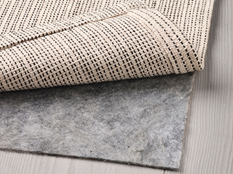 Detail of the back of a natural woven rug, showing its texture and non-slip reverse, layered over a gray felt pad for added comfort and stability.