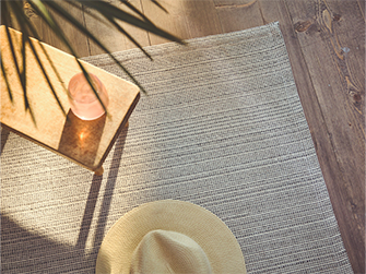Flat-woven rug in neutral tones with a wooden side table and a straw hat, under sunlight streaming through a window, creating soft shadows.