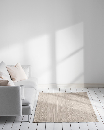 Natural woven rug in a minimalist living room with white walls and light wood flooring, in front of a simply designed sofa with soft cushions, enhanced by sunlight casting shadows.