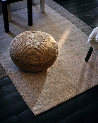 Natural fiber rug in a light beige tone with a textured wicker pouf in the center, and dark-legged furniture around, in a space with dark wood flooring and natural light.