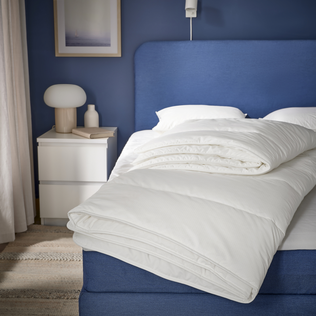 Modern bedroom with navy blue walls and a bed with a blue upholstered headboard, showing a folded white duvet on the bed and a white nightstand with a lamp and decorative items.